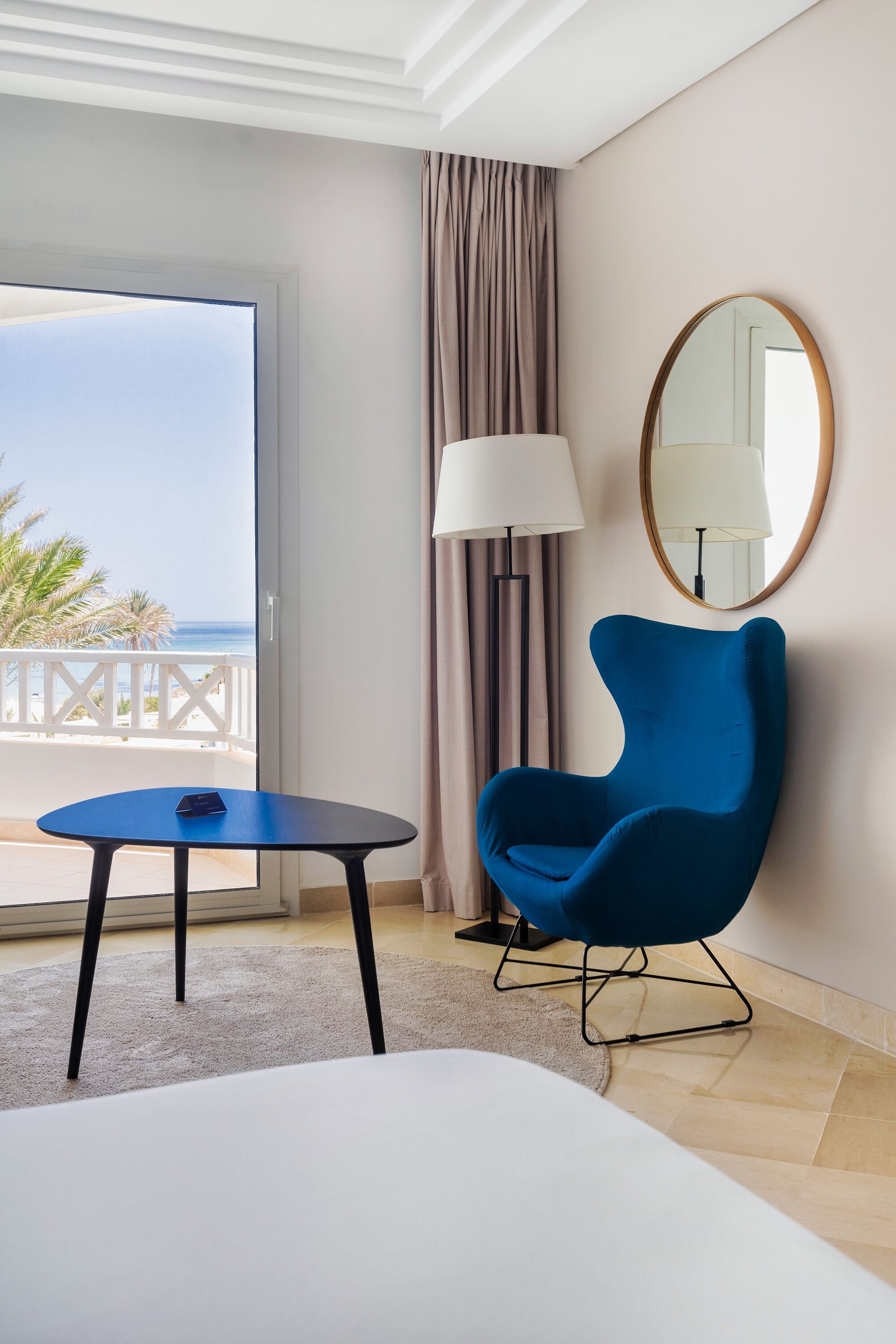 A hotel room with a balcony, a blue armchair, a small table, a lamp, and a large mirror.