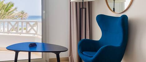 A hotel room with a balcony, a blue armchair, a small table, a lamp, and a large mirror.