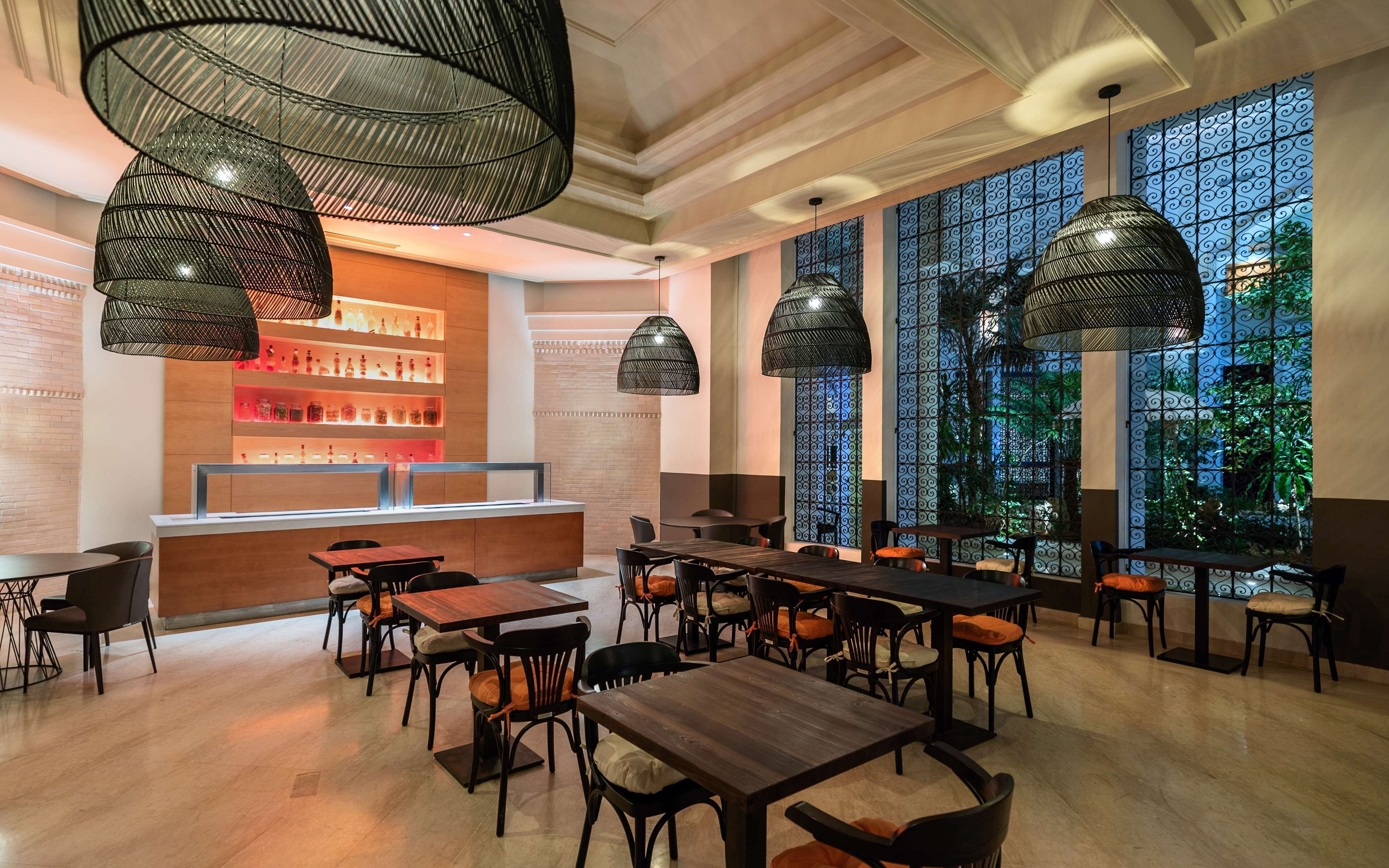A restaurant interior with high ceilings, black woven pendant lights, a bar area with shelves of bottles, and a view through a patterned window.