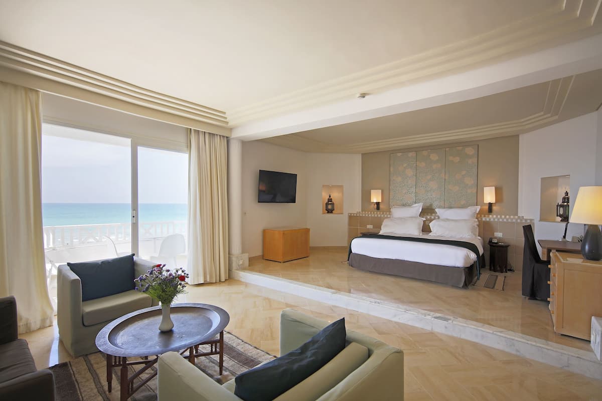 A hotel room with a bed, a desk, a chair, a small table with a vase, and a view of the ocean.