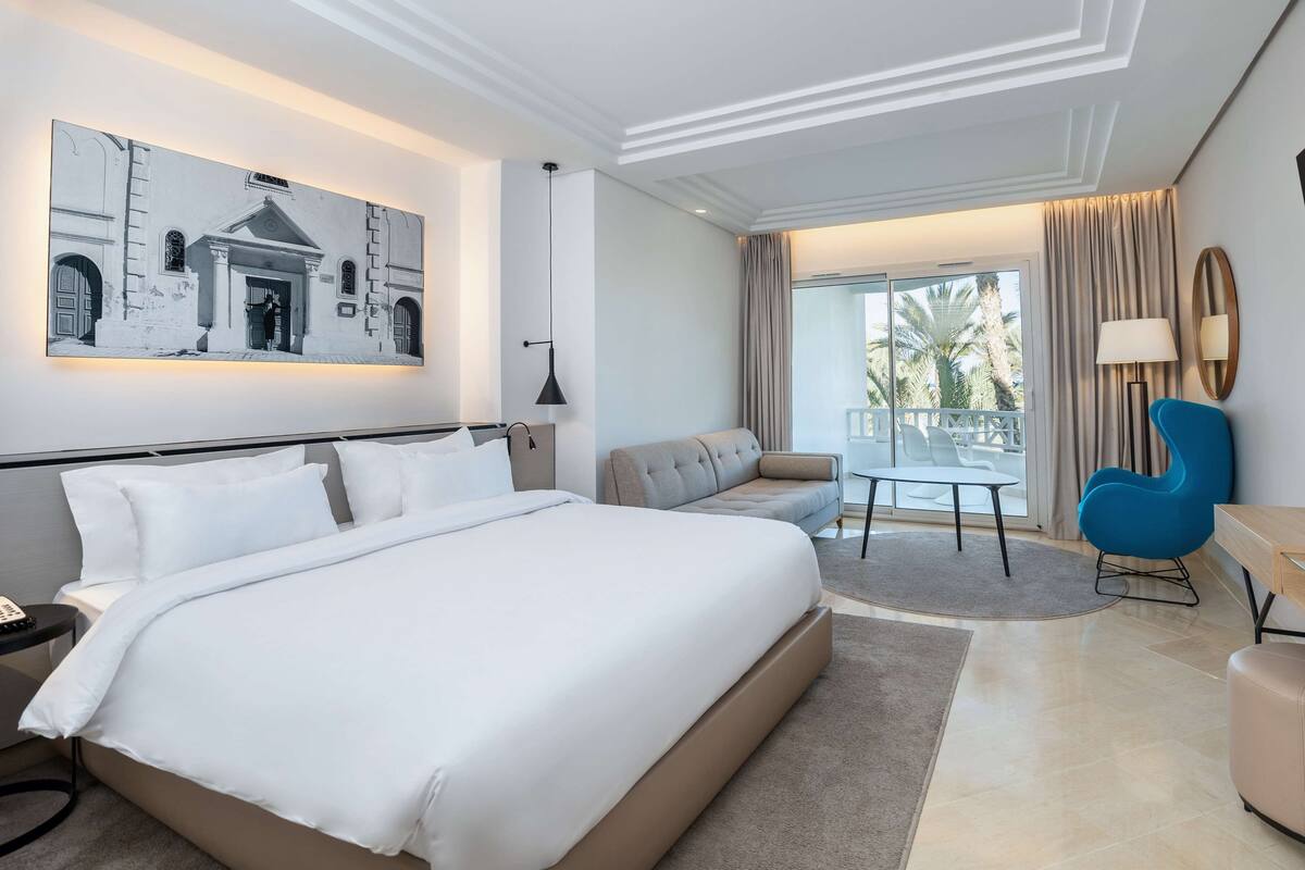 A modern hotel room with a large bed, a sofa, a blue armchair, and a balcony with a view of palm trees.