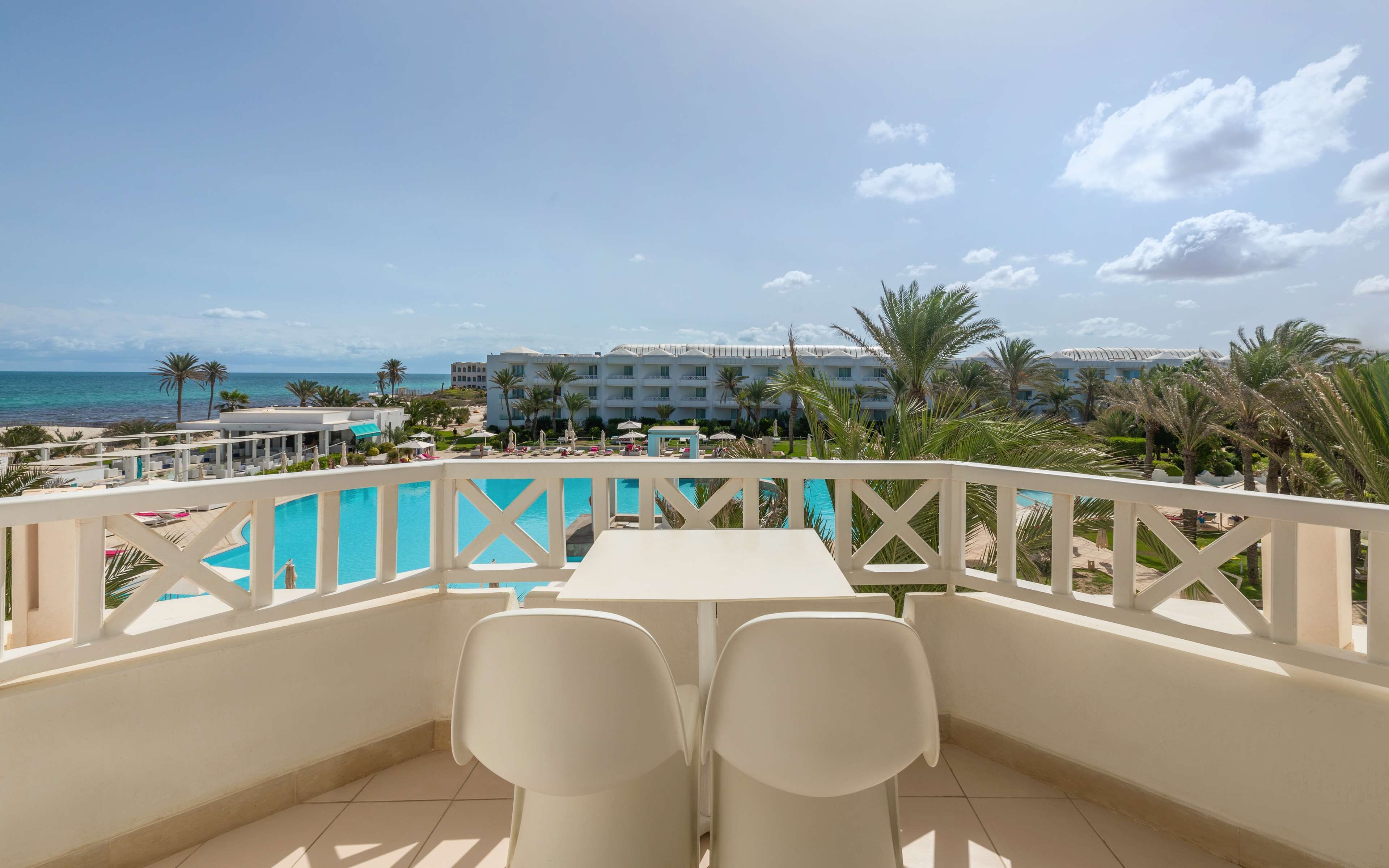 A balcony with two white chairs, a pool, and a view of the beach and hotel buildings.