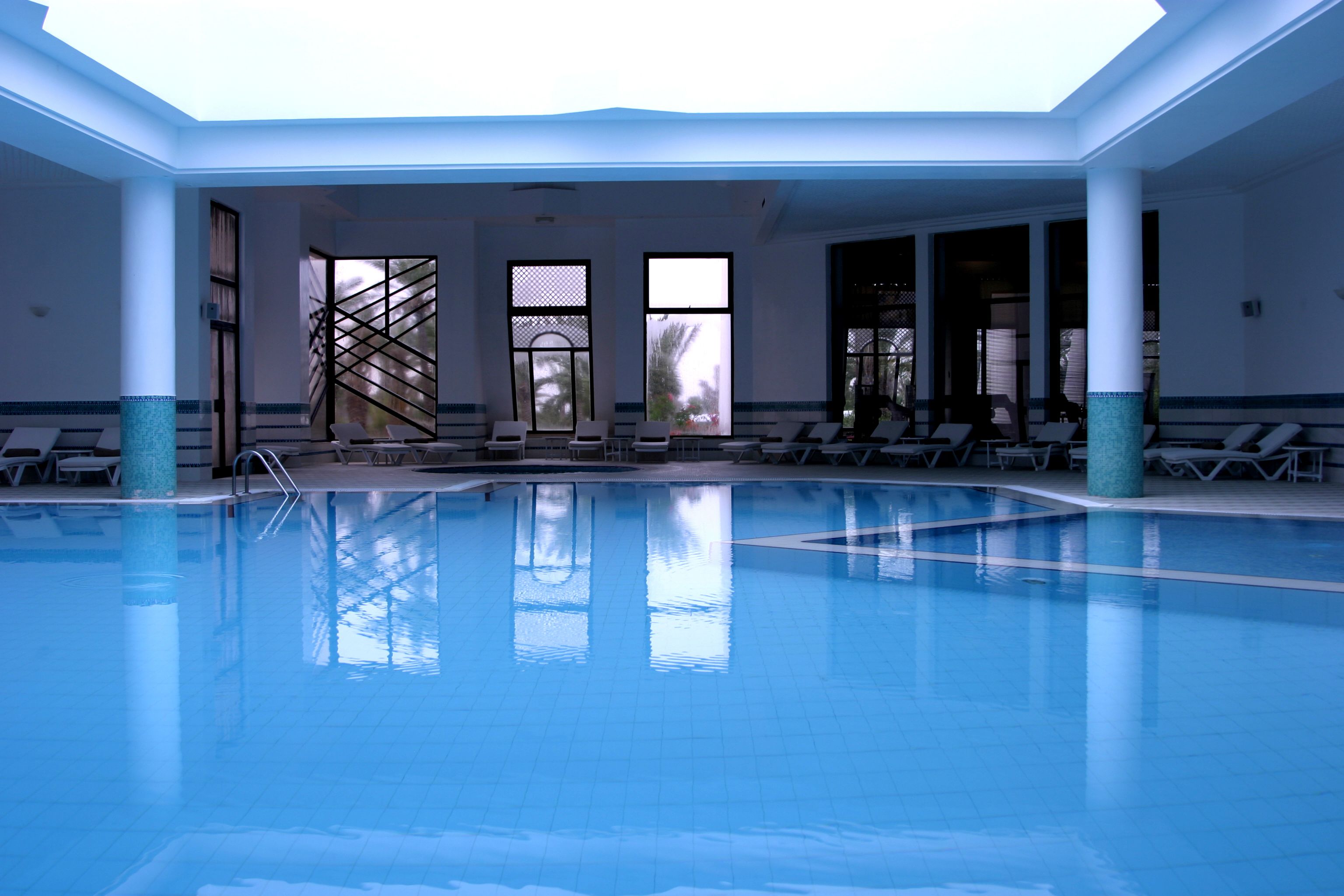 An indoor swimming pool with clear blue water, surrounded by lounge chairs and a covered area with large windows.