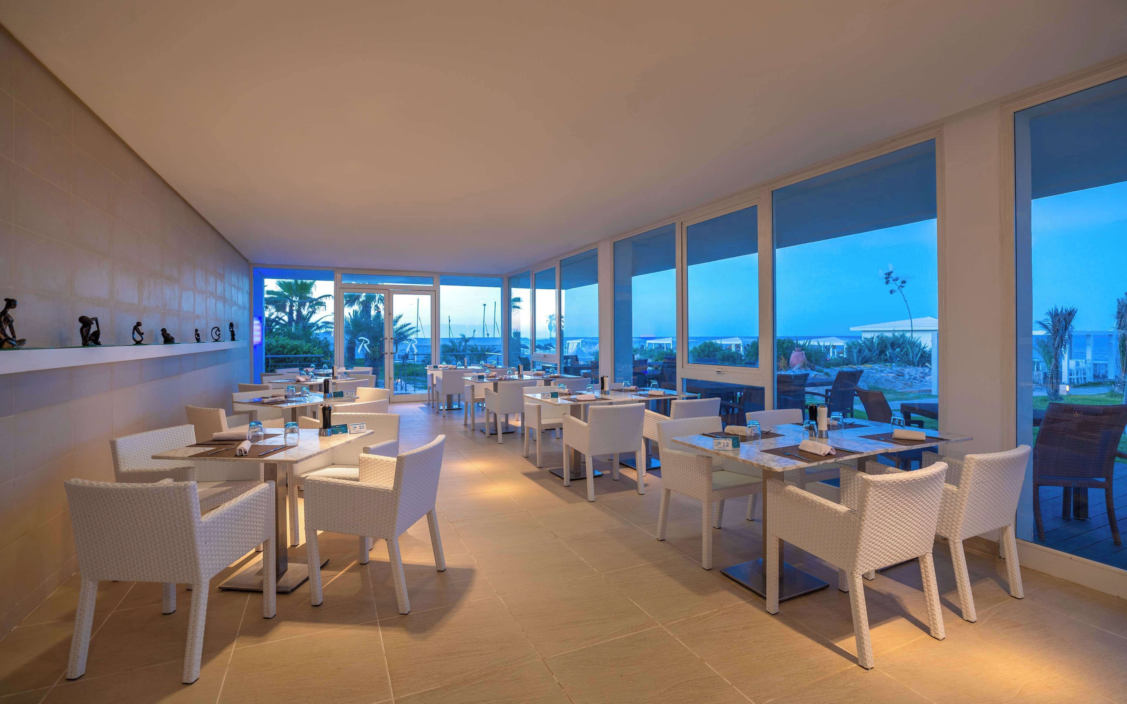 Un restaurante con mobiliario de mimbre blanco, ventanas amplias y vista al océano.