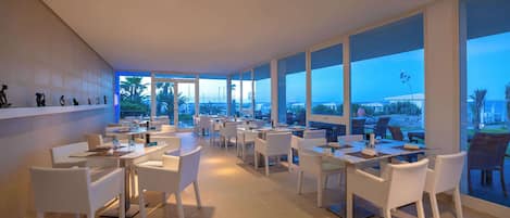 A restaurant with white wicker furniture, large windows, and a view of the ocean.