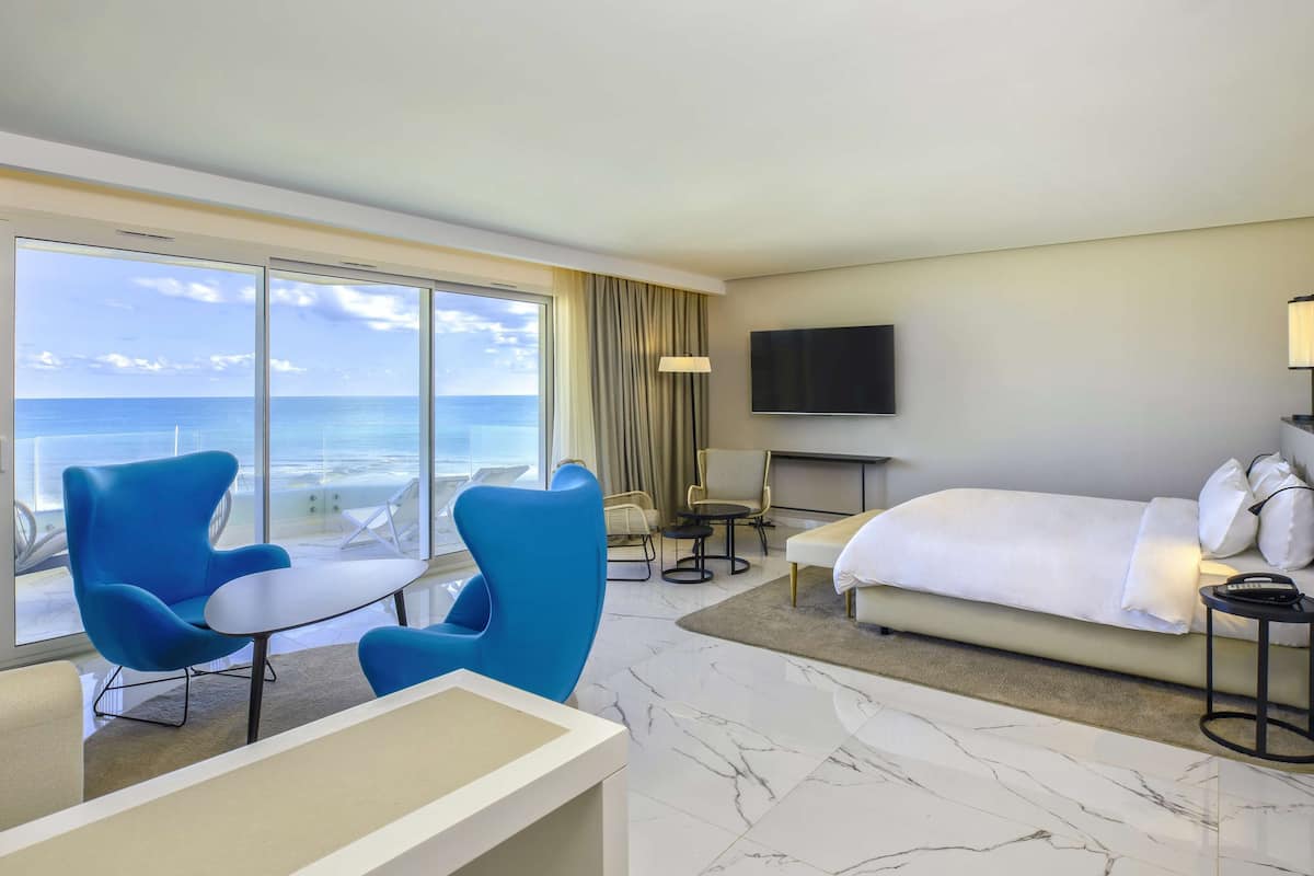 A hotel room with a large window offering a view of the ocean, a bed, a desk, two blue armchairs, and a flat-screen TV.