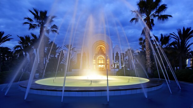 A fountain with water jets in a circular pool, surrounded by palm trees, with a building in the background. - Radisson Blu Athénée Palace Resort & Thalasso, Djerba (Mezraia)