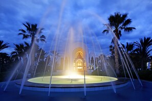 Una fuente con chorros de agua en un estanque circular, rodeada de palmeras, con un edificio al fondo.