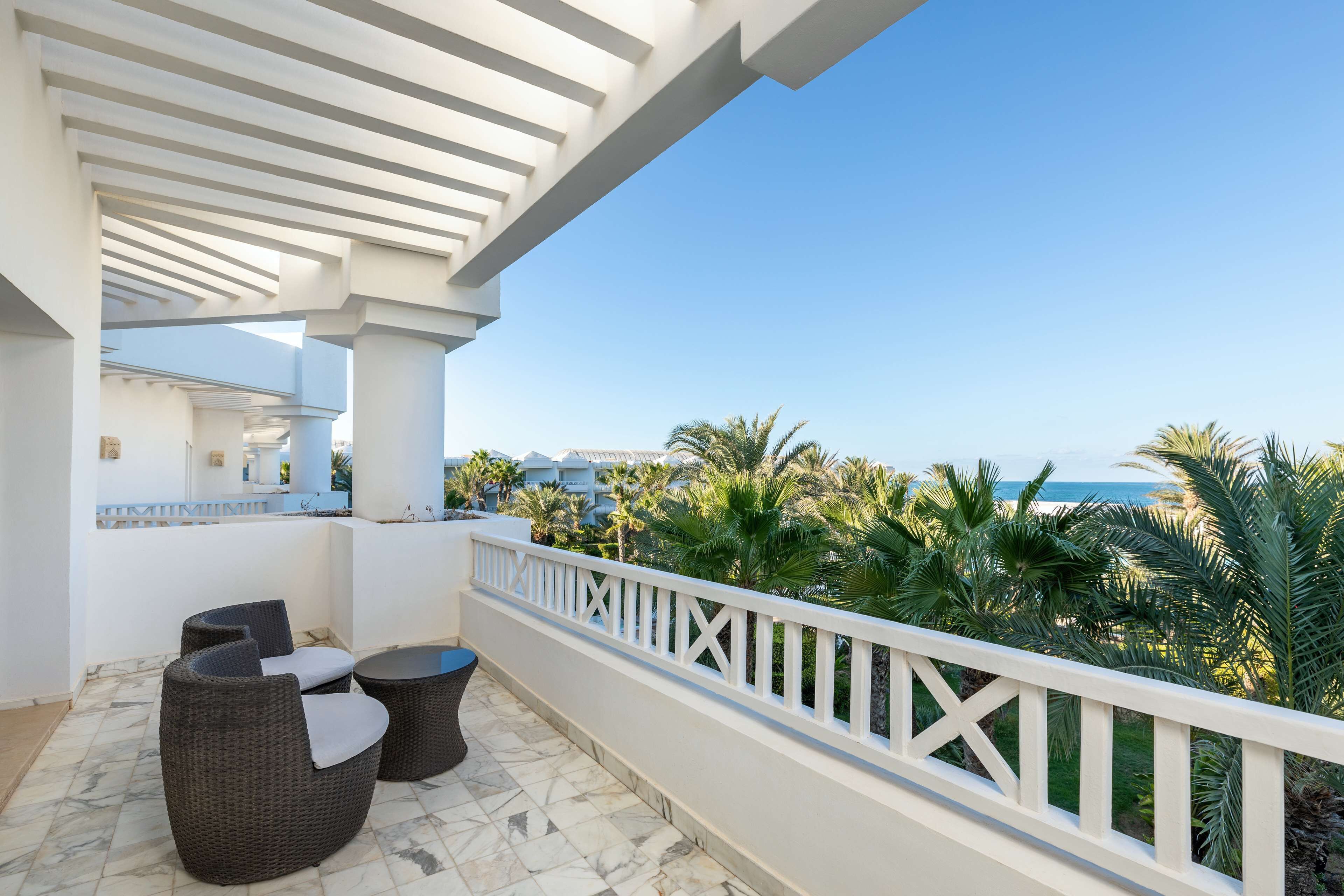 Un balcon doté de meubles en osier, d’une balustrade blanche et offrant une vue sur les palmiers et la mer.