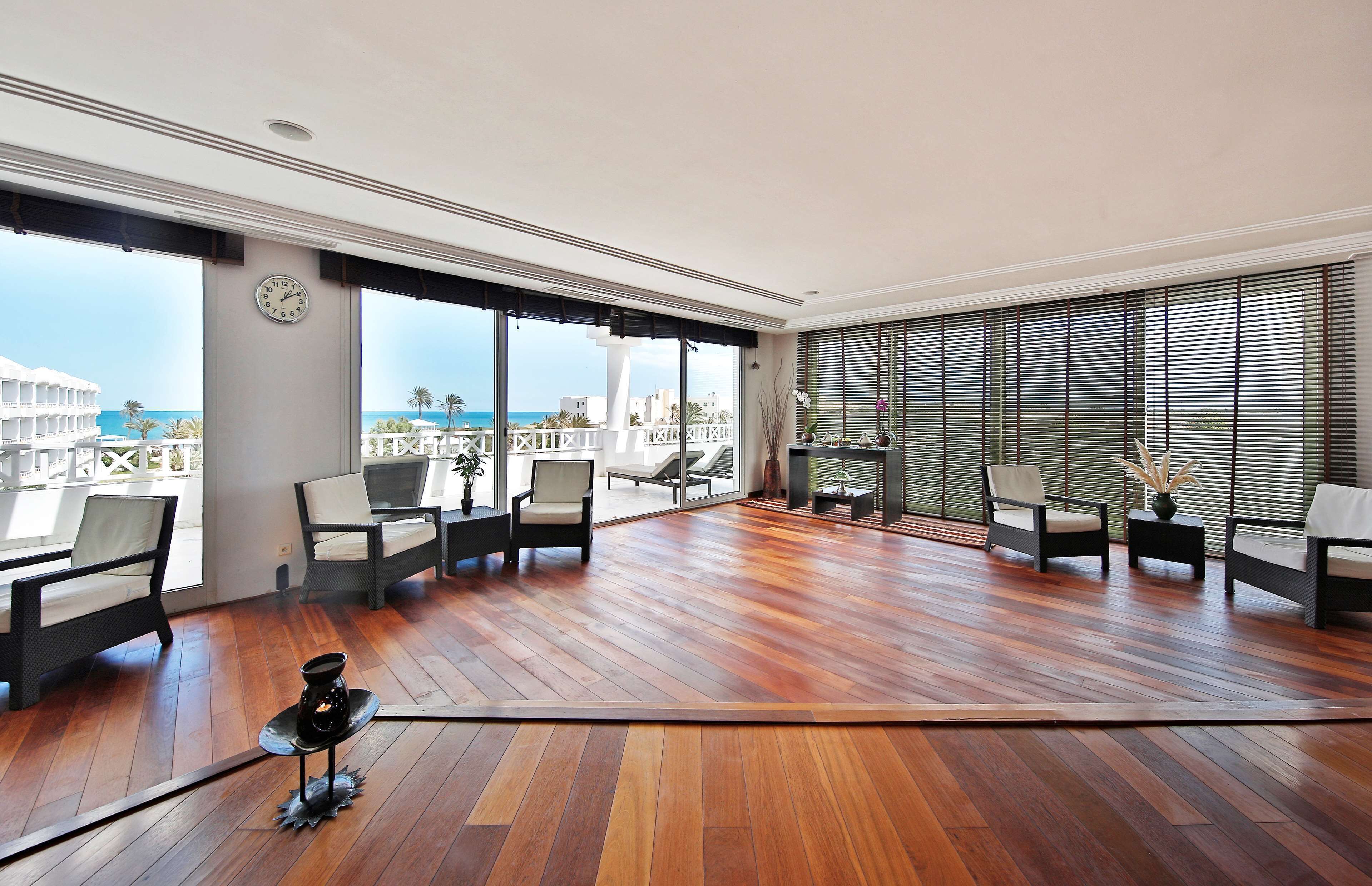 A spacious balcony with wooden flooring, multiple seating areas, and large windows offering a view of the sea and buildings.