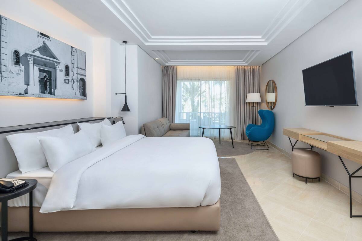 A modern hotel room with a large bed, a desk, a chair, a TV, and a painting on the wall.
