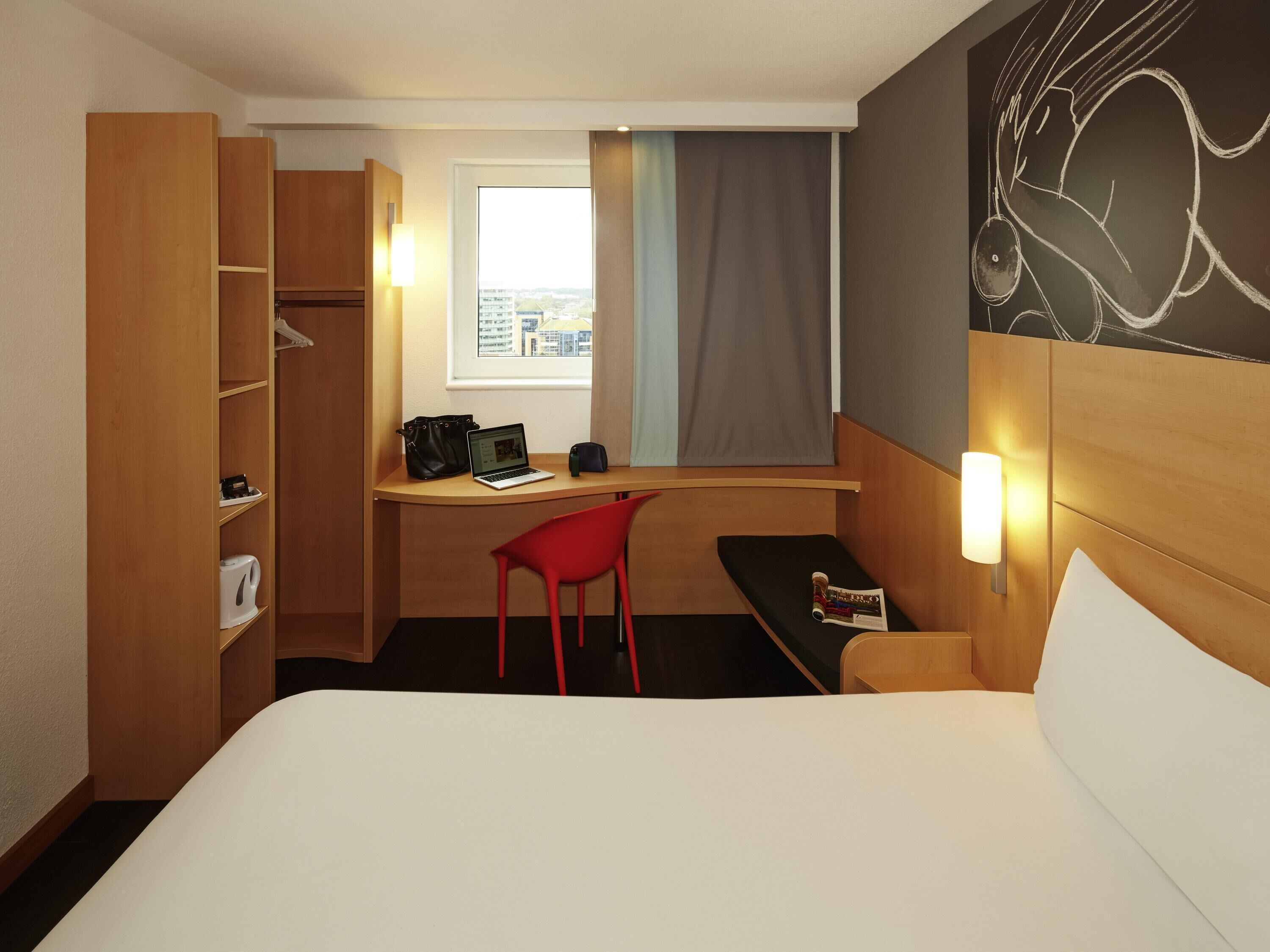 Habitación de hotel con cama, escritorio con una computadora portátil, una silla roja, ventana con cortinas y pared con diseño de pizarra.