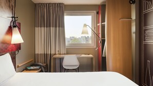 Een hotelkamer met een bed, een bureau met stoel, een lamp, een raam met gordijnen en uitzicht op de stad.