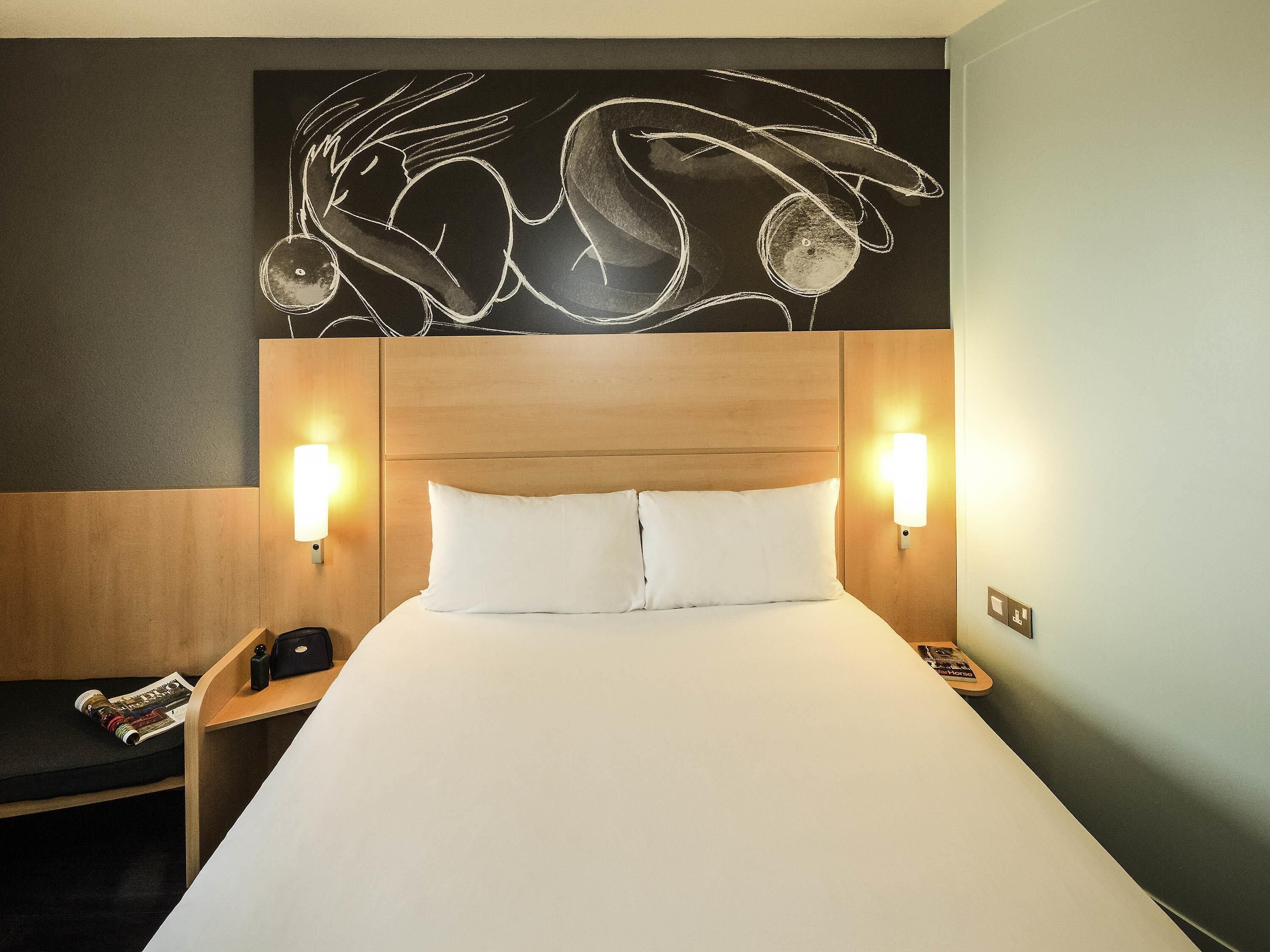 A hotel room with a wooden headboard, two bedside lamps, a bed with white linens, and a wall art piece above the bed.