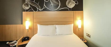 Una habitación de hotel con cabecera de madera, dos lámparas de noche, una cama con ropa blanca y una obra de arte en la pared sobre la cama.