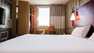 A hotel room with a large bed, a desk with a lamp, a window with curtains, and a wall with a decorative panel.