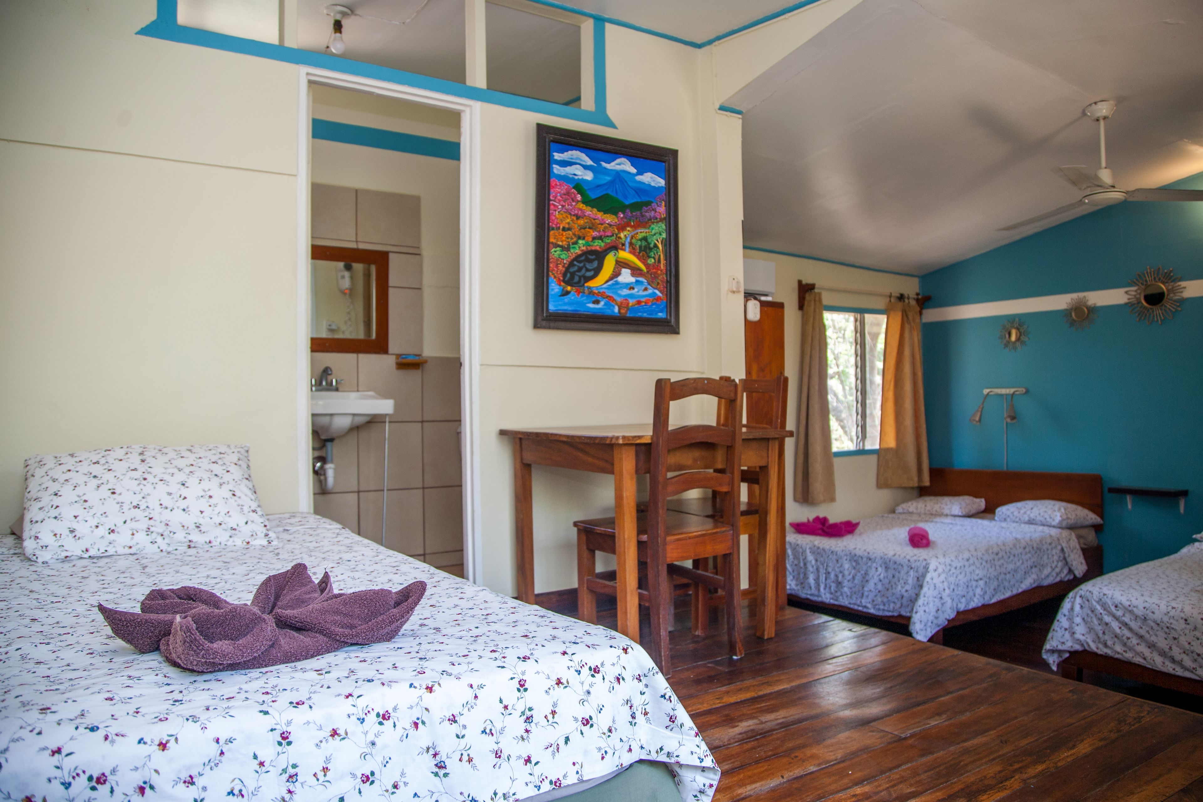 Une chambre avec deux lits, une chaise en bois, un tableau et une salle de bain visible à travers une porte ouverte.