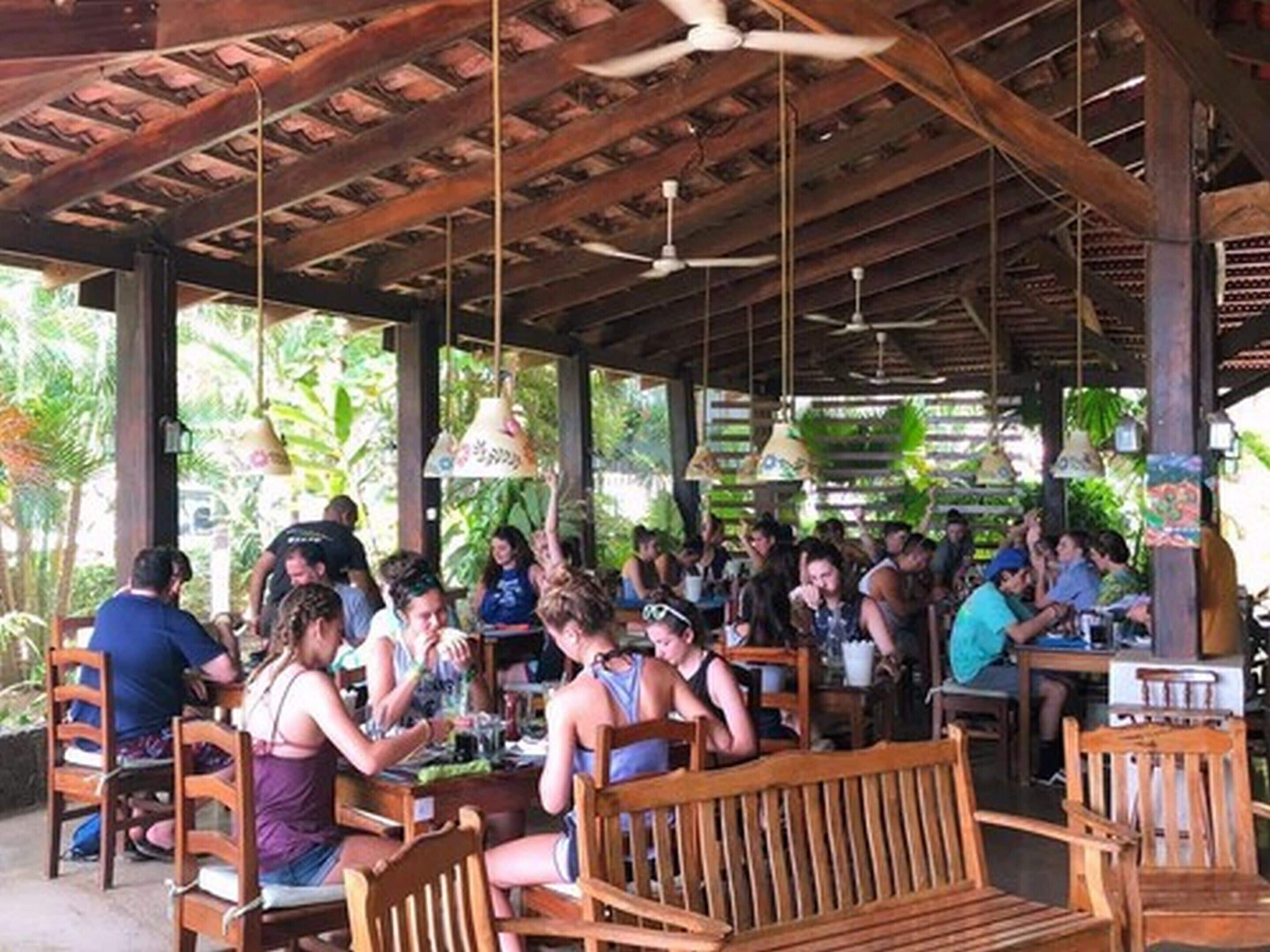 An outdoor dining area with wooden tables and chairs, covered by a wooden roof with ceiling fans.