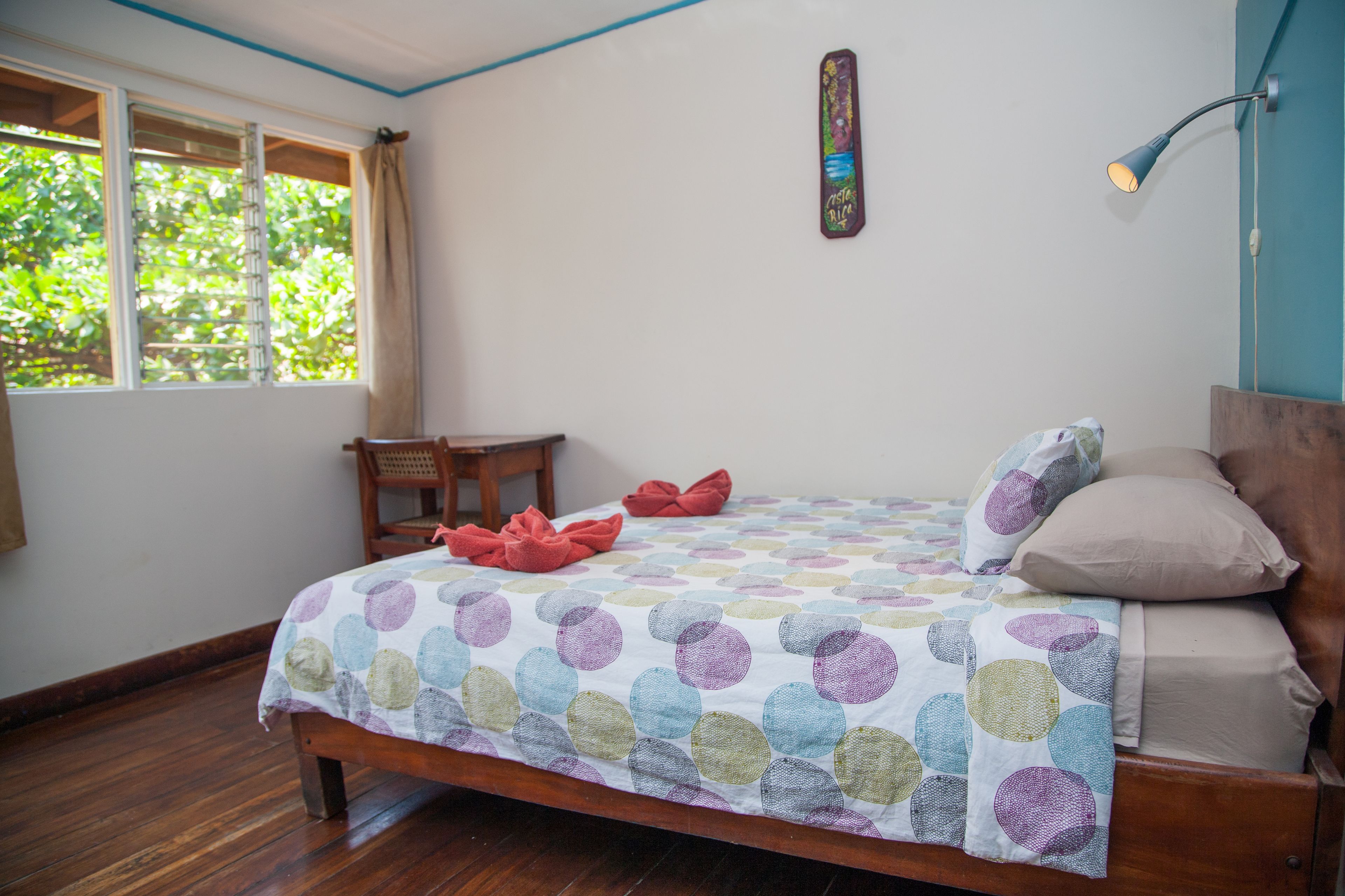 Une chambre à coucher avec un lit en bois, une couvre-lit à pois colorés, une petite chaise en bois, une table de chevet et une fenêtre avec des rideaux.