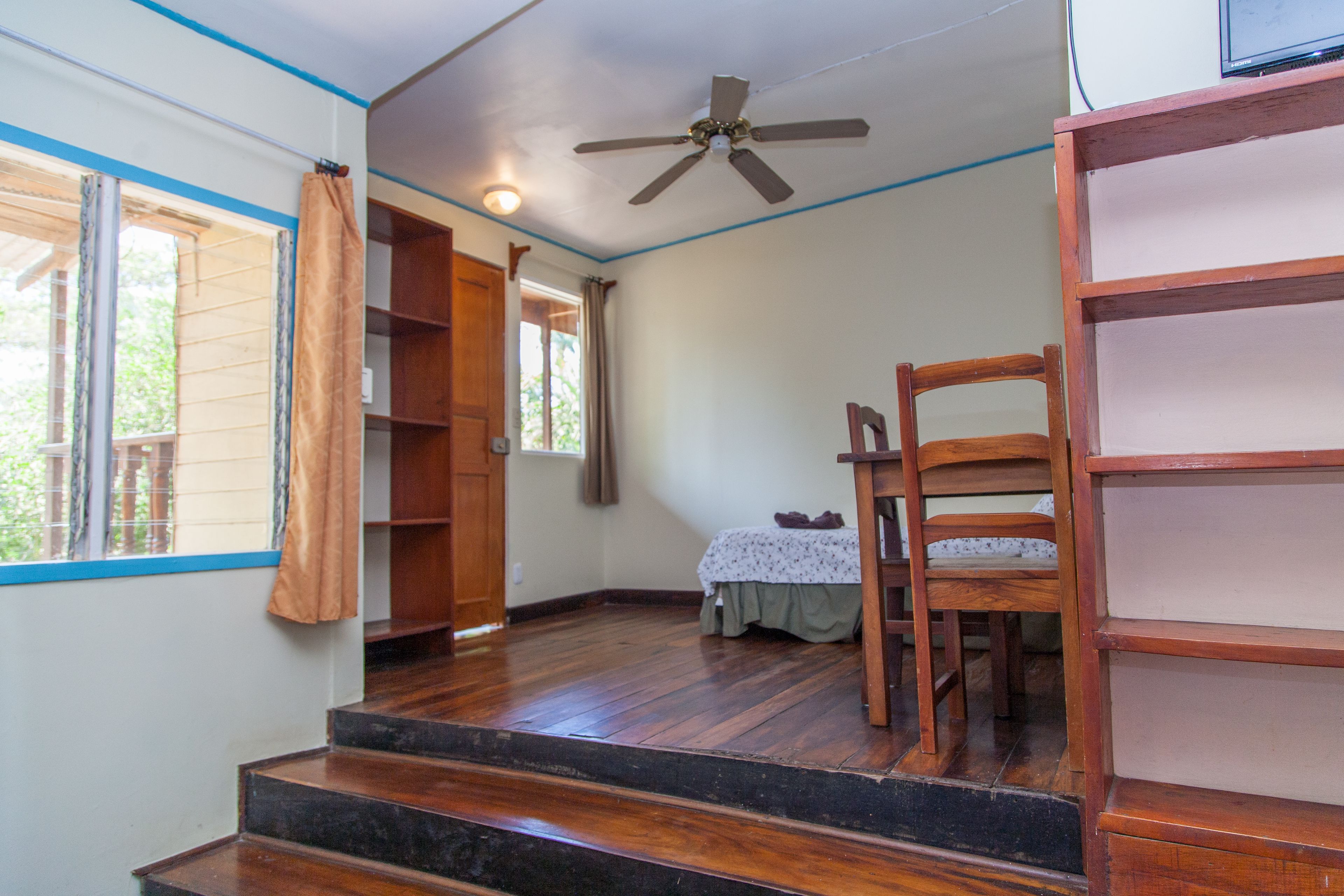 Une pièce avec un plancher en bois, un ventilateur de plafond, un lit recouvert d’une courtepointe à motifs floraux et une chaise en bois.