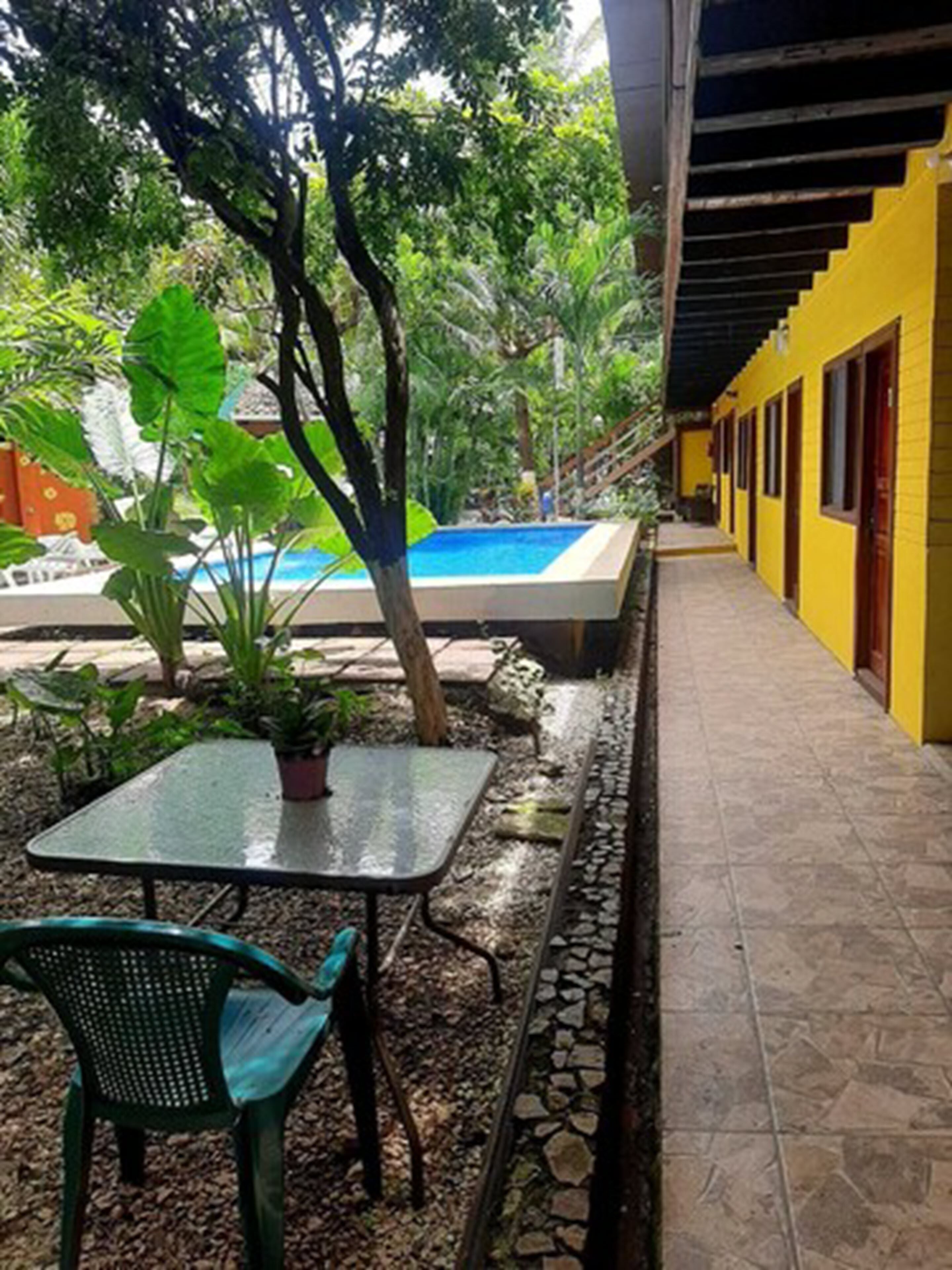 A covered outdoor area with a pool, a table with chairs, and a yellow building in the background.