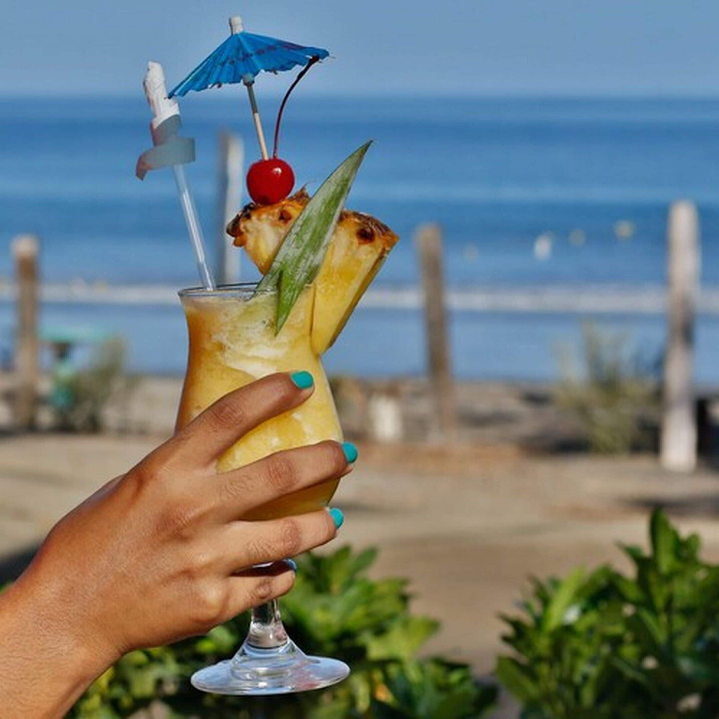 Una mano sostiene un cóctel con una cereza y una rodaja de piña, con un parasol azul y una pajita, frente a un paisaje de playa.