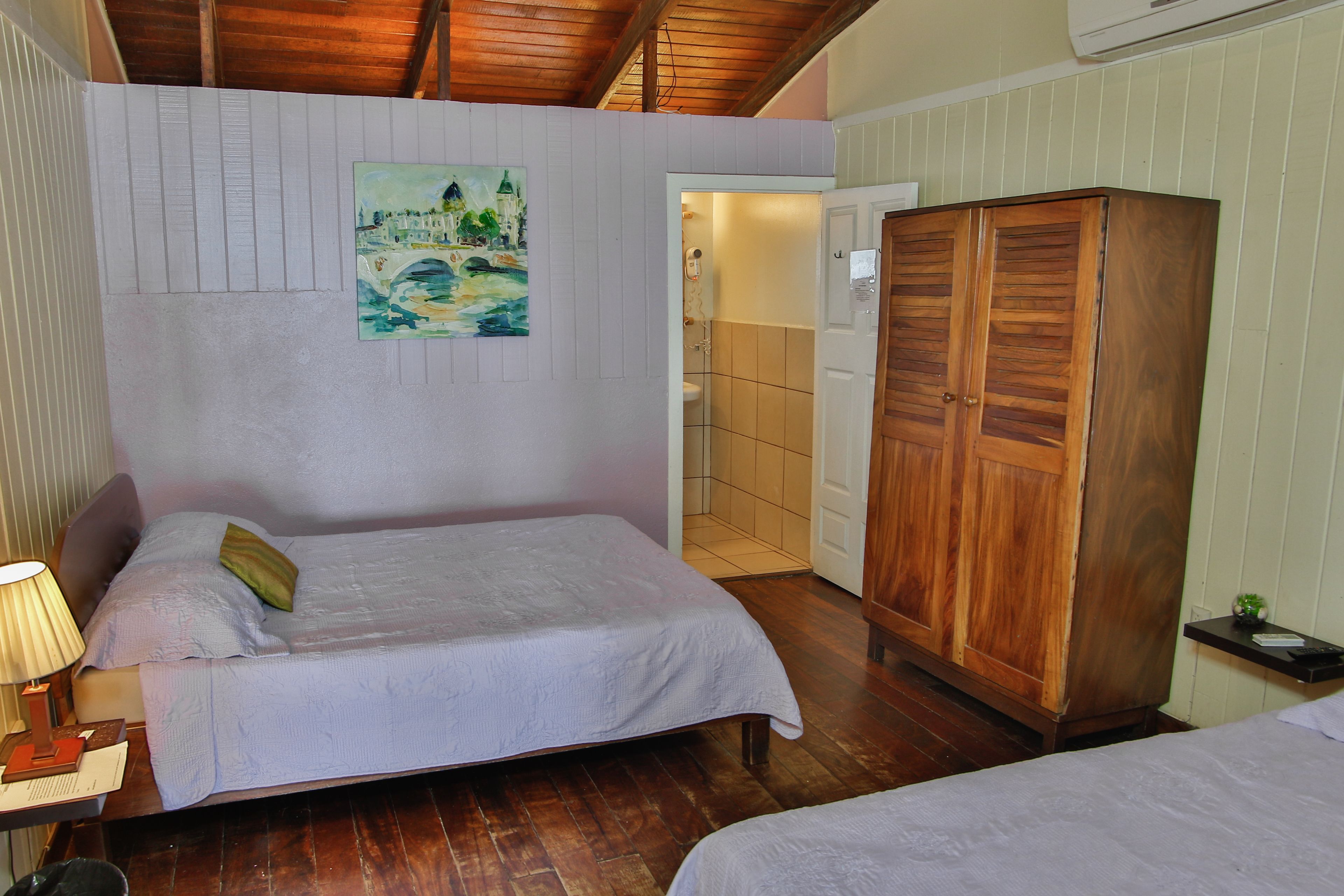 Un dormitorio con dos camas, un armario de madera, un cuadro en la pared y un baño visible a través de una puerta abierta.