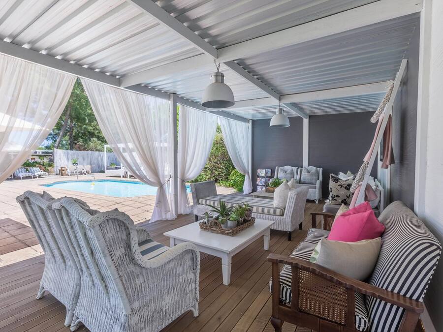 A covered outdoor seating area with wicker furniture, a white table, and a pool in the background.