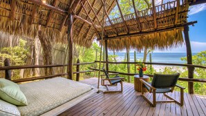 Une terrasse en bois avec un toit de chaume, un canapé et deux fauteuils donnant sur une plage.