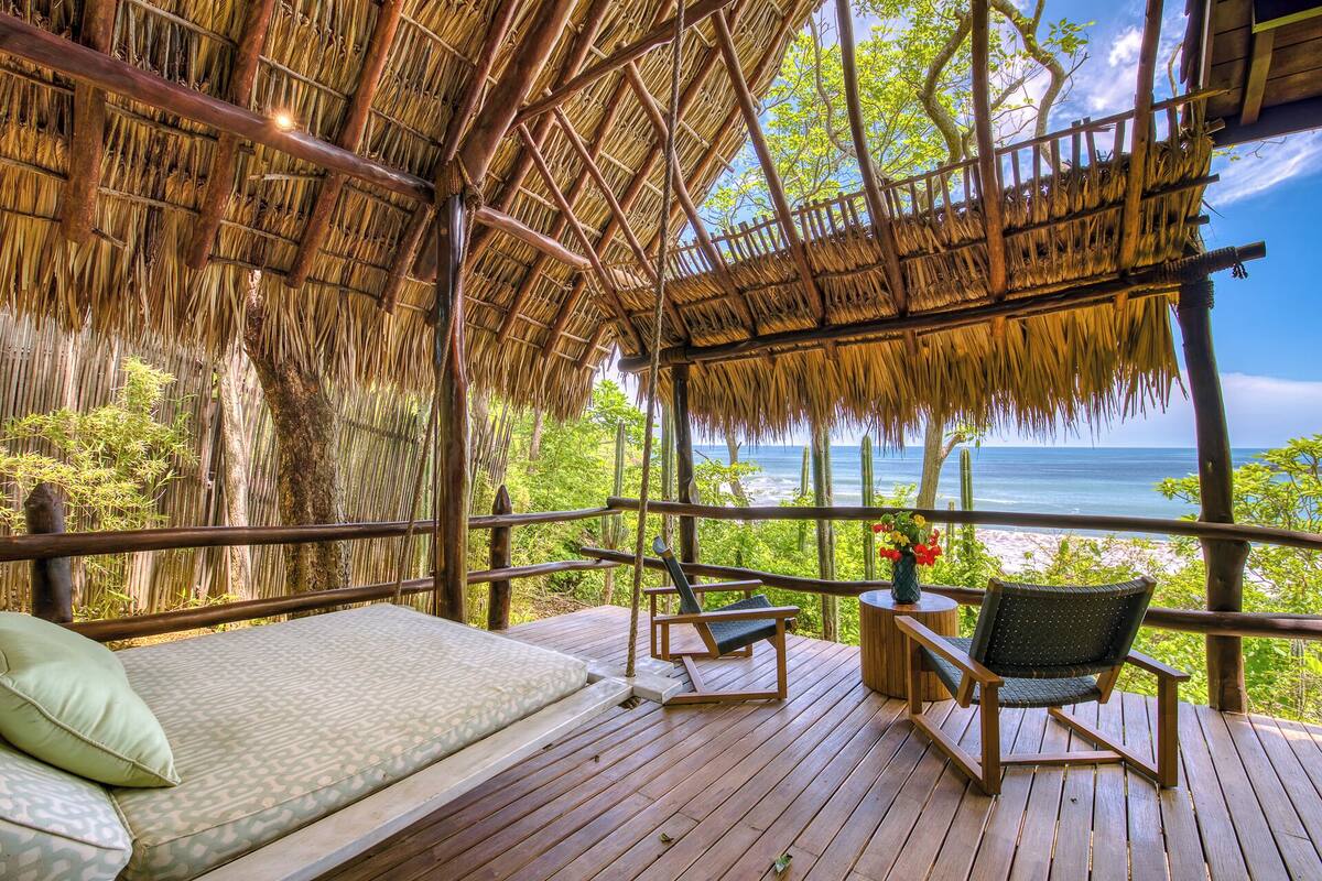 Una terraza de madera con techo de paja, un sofá y dos sillas con vistas a una playa.