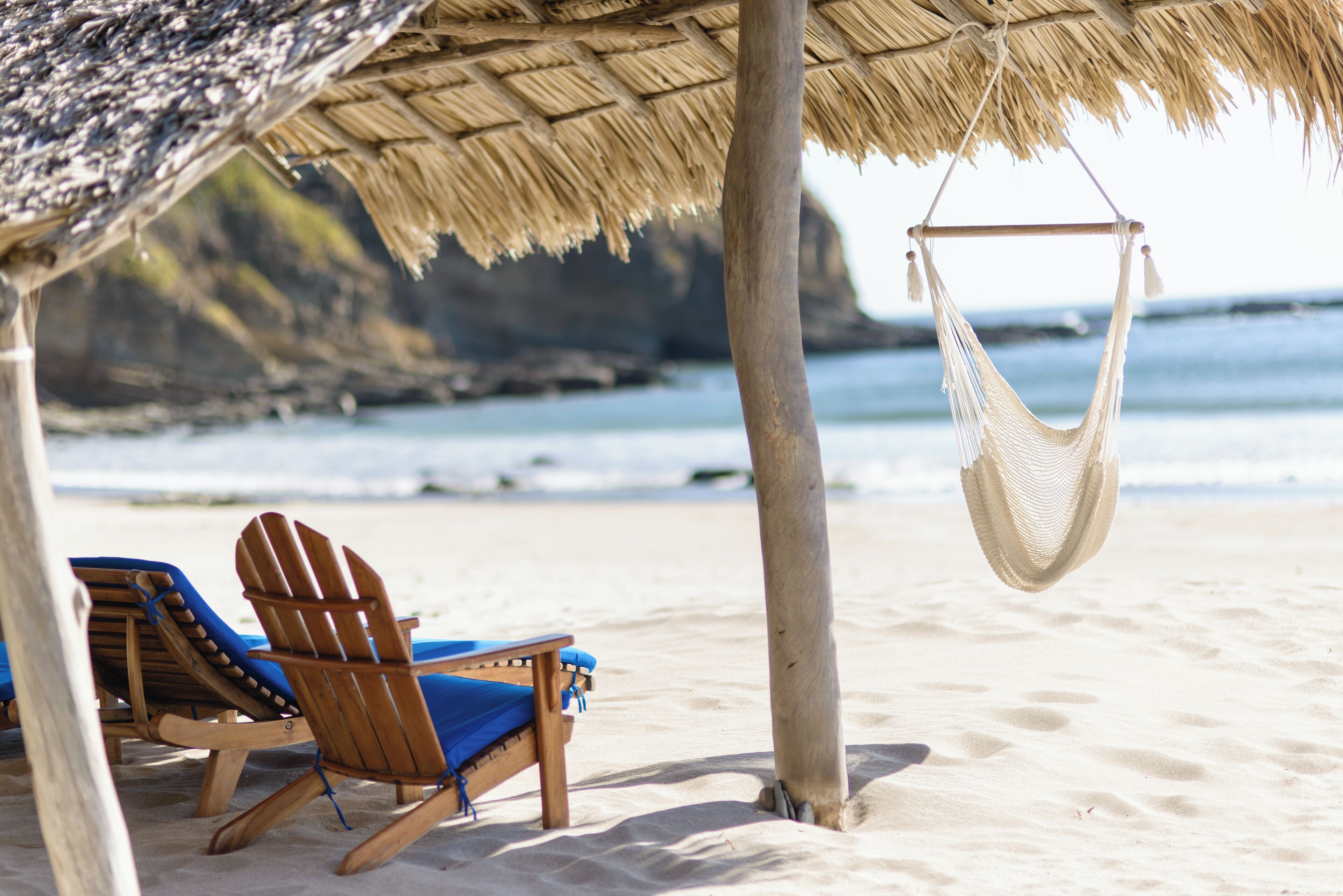 A beach scene with wooden chairs and a hammock under a thatched roof.