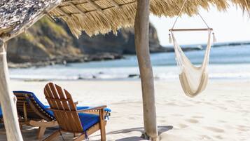 A beach scene with wooden chairs and a hammock under a thatched roof.