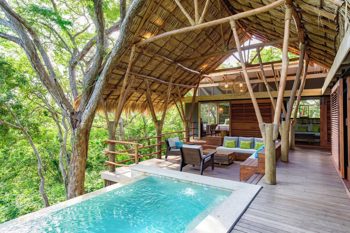 Zona de piscina exterior con terraza de madera, sillas de descanso y una estructura cubierta de paja.
