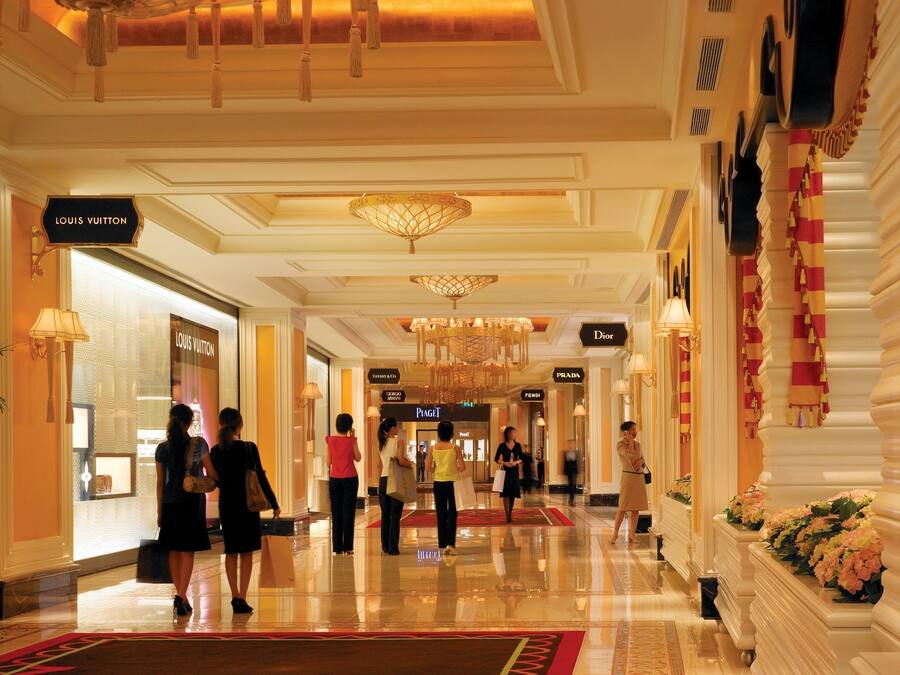 Un pasillo de hotel con señalización de marcas de lujo, incluyendo Louis Vuitton y Dior, y un piso de mármol pulido.