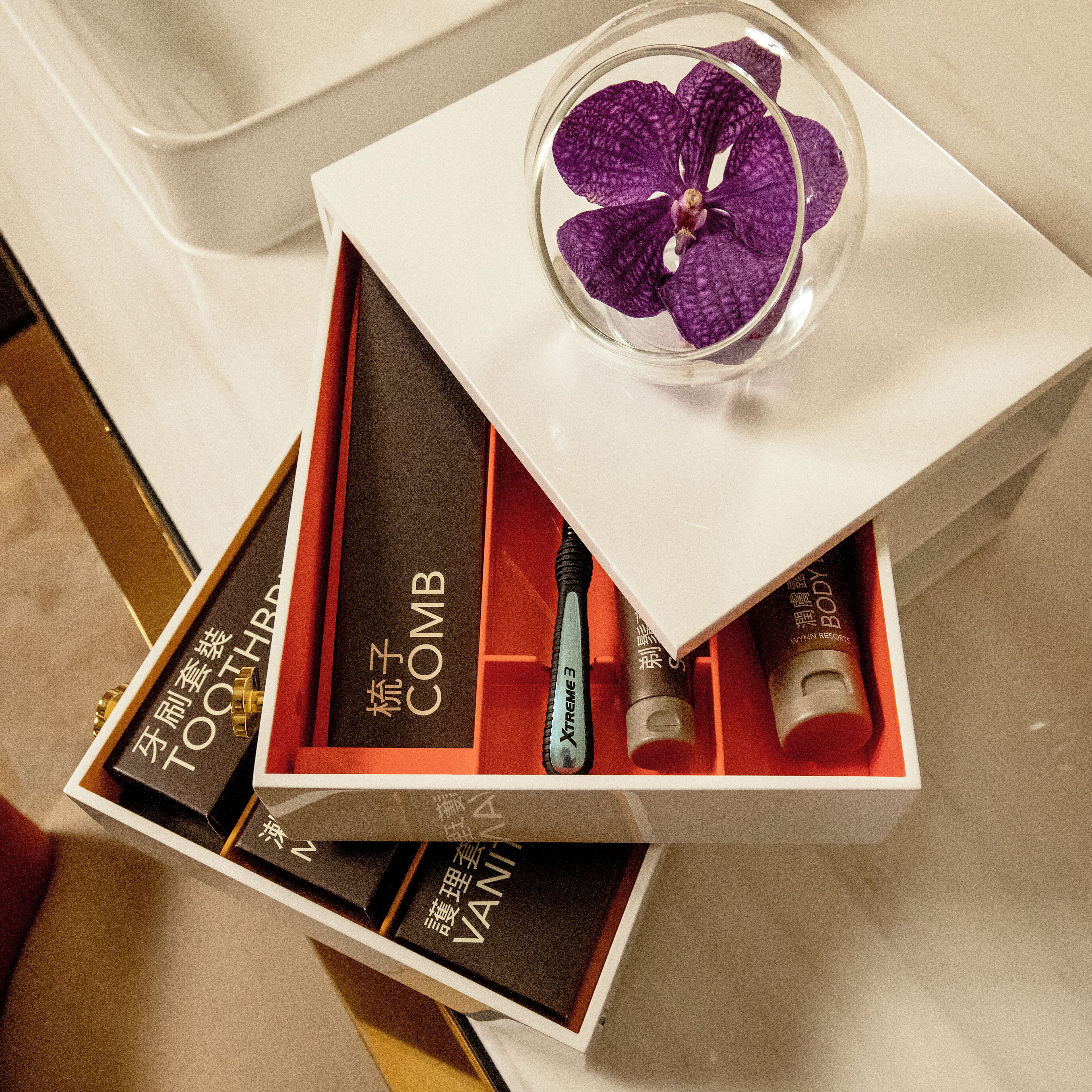 A white box with a purple orchid, containing a toothbrush and two tubes labeled 'COMB' and 'VANISH'.
