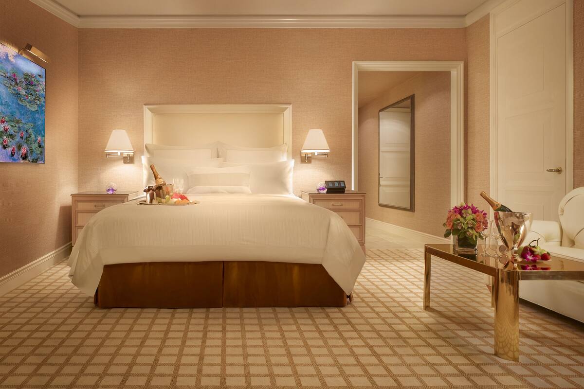 Habitación de hotel con una cama grande, dos mesitas de noche, un pequeño sillón blanco, una mesa con un florero y un cuadro en la pared.