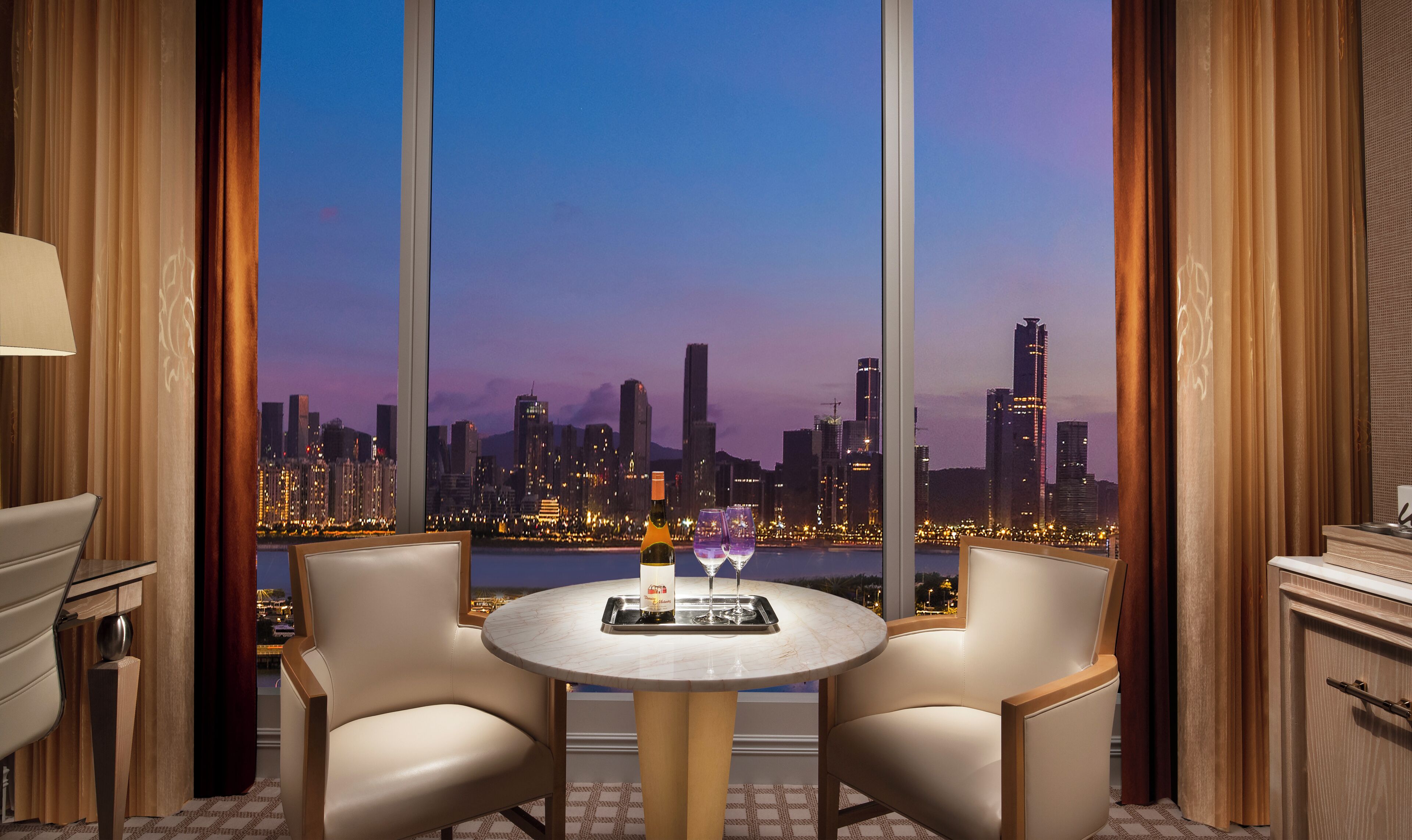 Una habitación con vista a un horizonte urbano al atardecer, una mesa redonda con dos sillas y una botella de vino sobre la mesa.