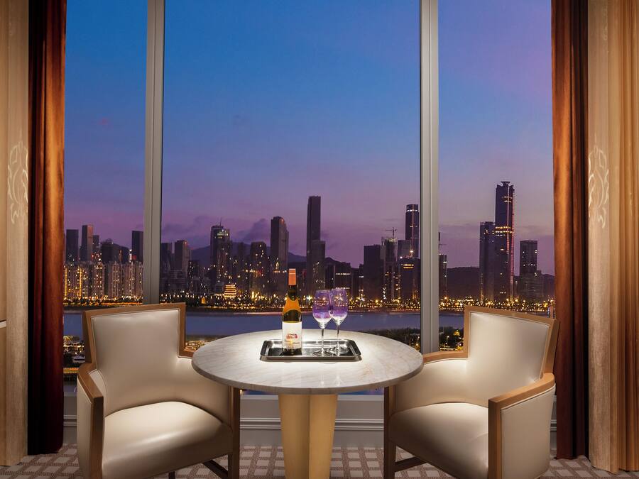 Una habitación con vista a un horizonte urbano al atardecer, una mesa redonda con dos sillas y una botella de vino sobre la mesa.