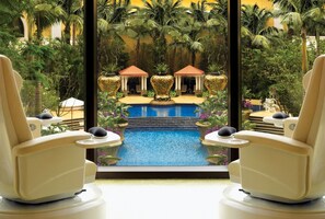 A luxurious indoor pool area with two reclining chairs facing a central pool, surrounded by tropical plants and a view of a building with red-roofed structures. - Wynn Macau (Macau)