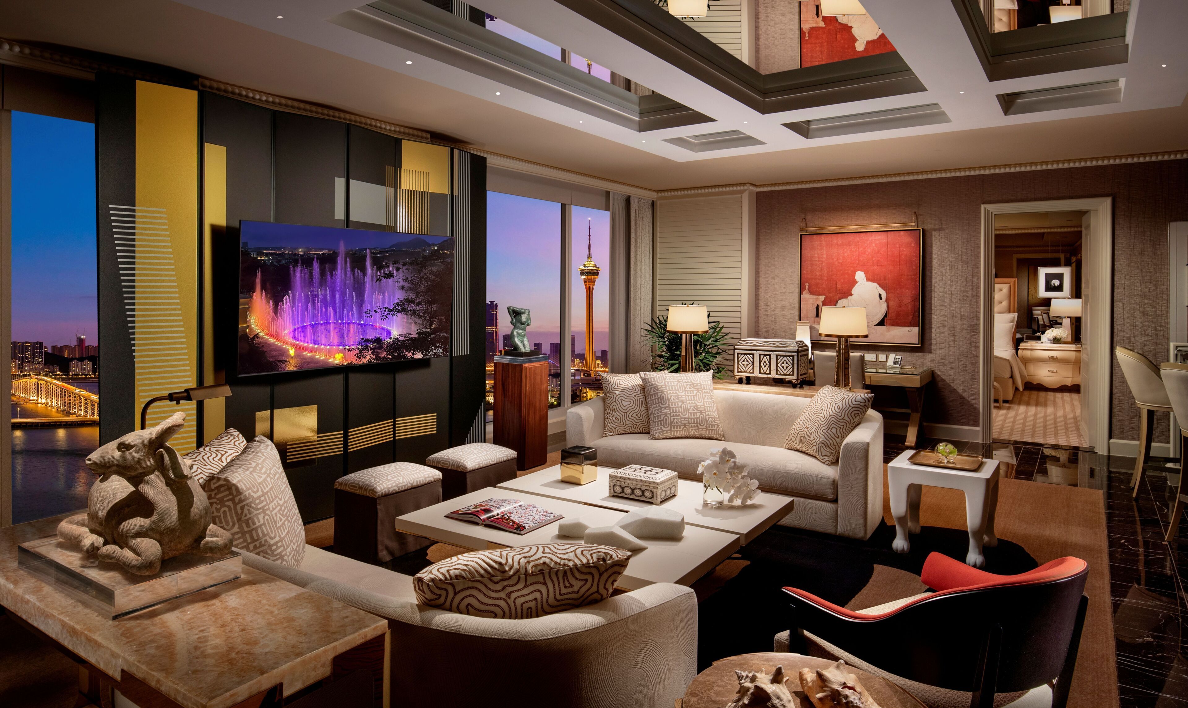 Habitación de hotel moderna con televisor grande, asientos cómodos, zona de comedor y vistas a la ciudad.