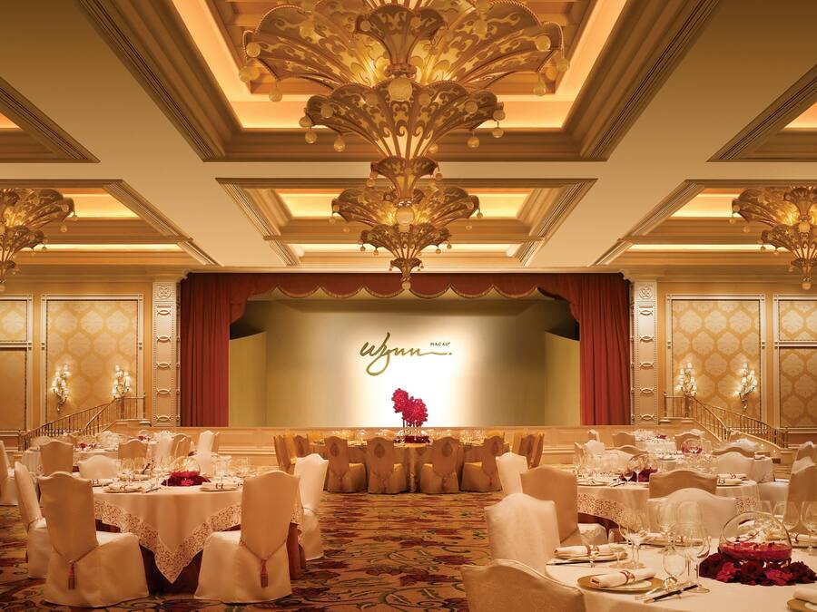 Une salle de banquet avec des tables rondes, des chaises blanches et une scène sur laquelle on peut lire l’inscription « W JUM MACAU ».