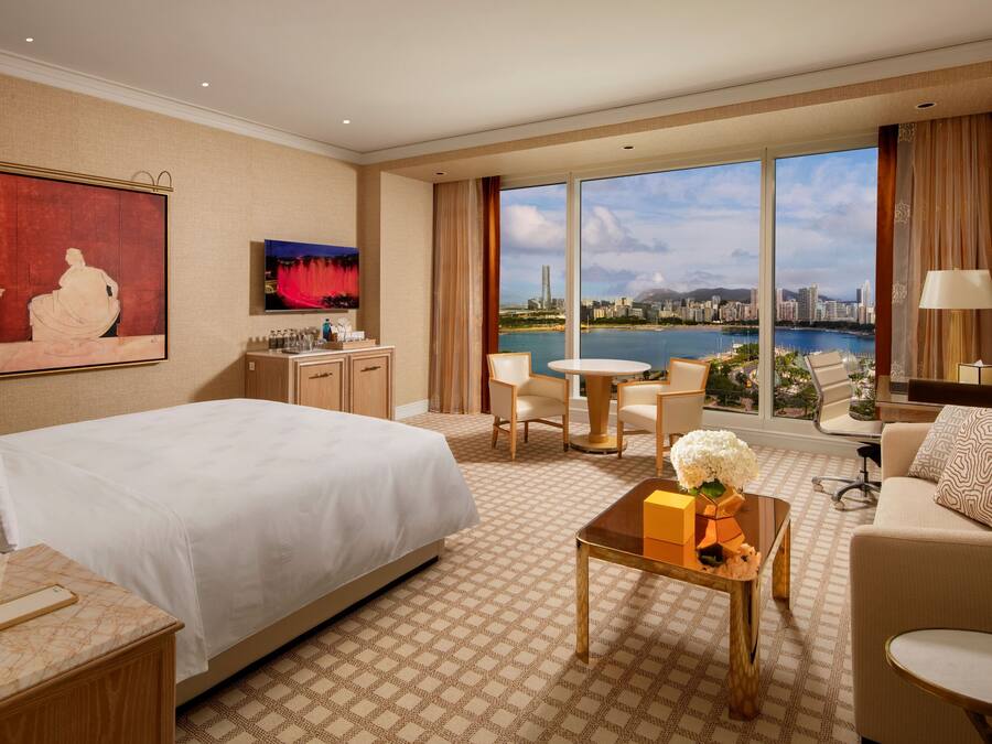 Habitación de hotel con una cama grande, un escritorio, una silla, una mesita y vistas a un horizonte urbano.
