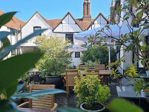 An outdoor seating area with tables and chairs, surrounded by potted plants and a building with a tiled roof. - The Kings Arms Hotel (Amersham)