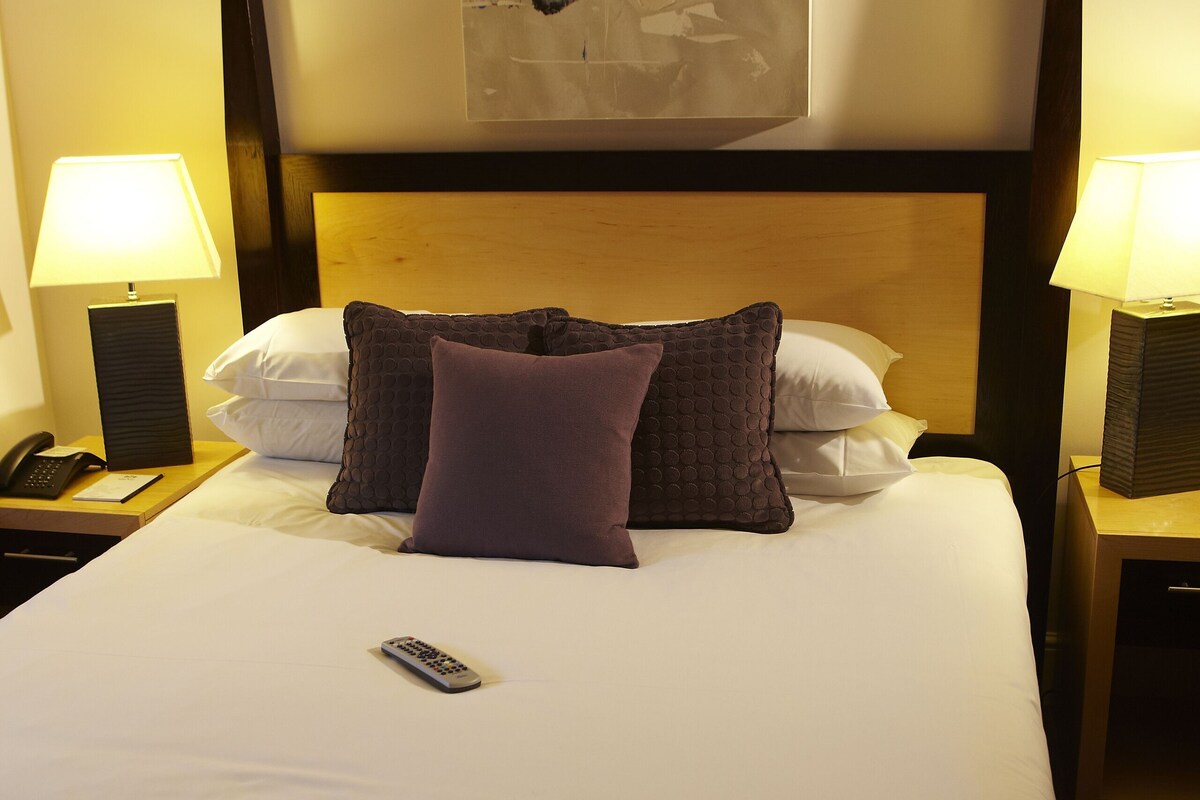 Una habitación de hotel con una cama, dos lámparas de noche, un control remoto y una obra de arte colgada en la pared.