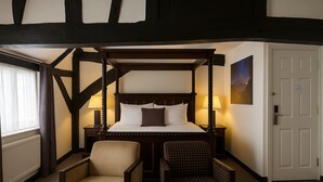 A four-poster bed with a headboard, two bedside tables with lamps, a chair, and a checkered ottoman. - The Kings Arms Hotel (Amersham)