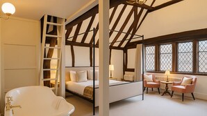 A bedroom with a large bathtub, a bed with white linens, a wooden ladder, and a window with curtains. - The Kings Arms Hotel (Amersham)