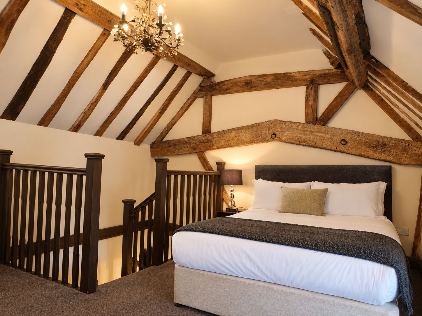 A bedroom with a sloped ceiling, exposed wooden beams, a bed with white linens, a dark headboard, and a chandelier.