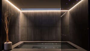 Un moderno spa con un jacuzzi rectangular, paredes de textura oscura y un florero con ramas secas.