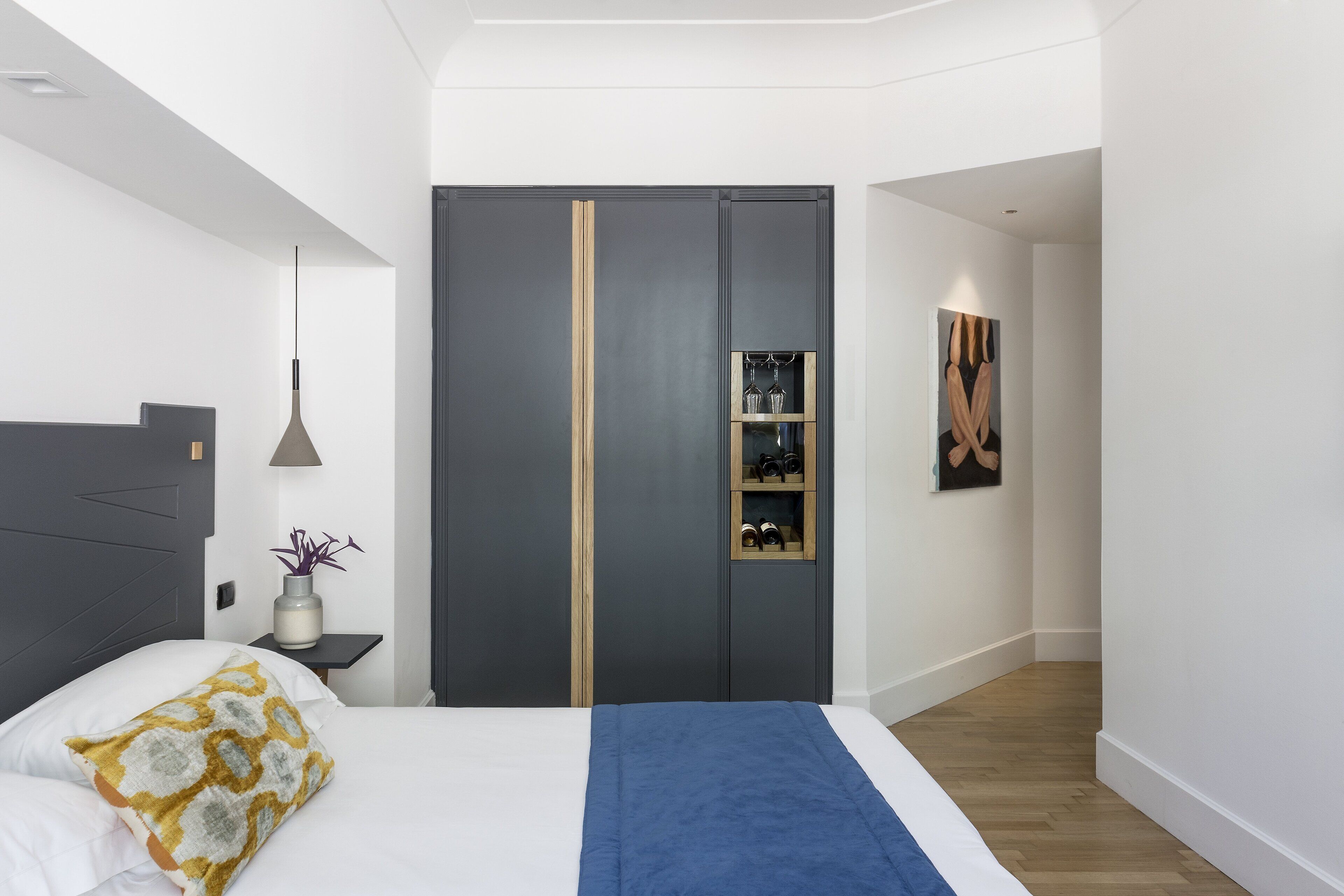 A modern bedroom with a bed, bedside table, lamp, wardrobe, and artwork.