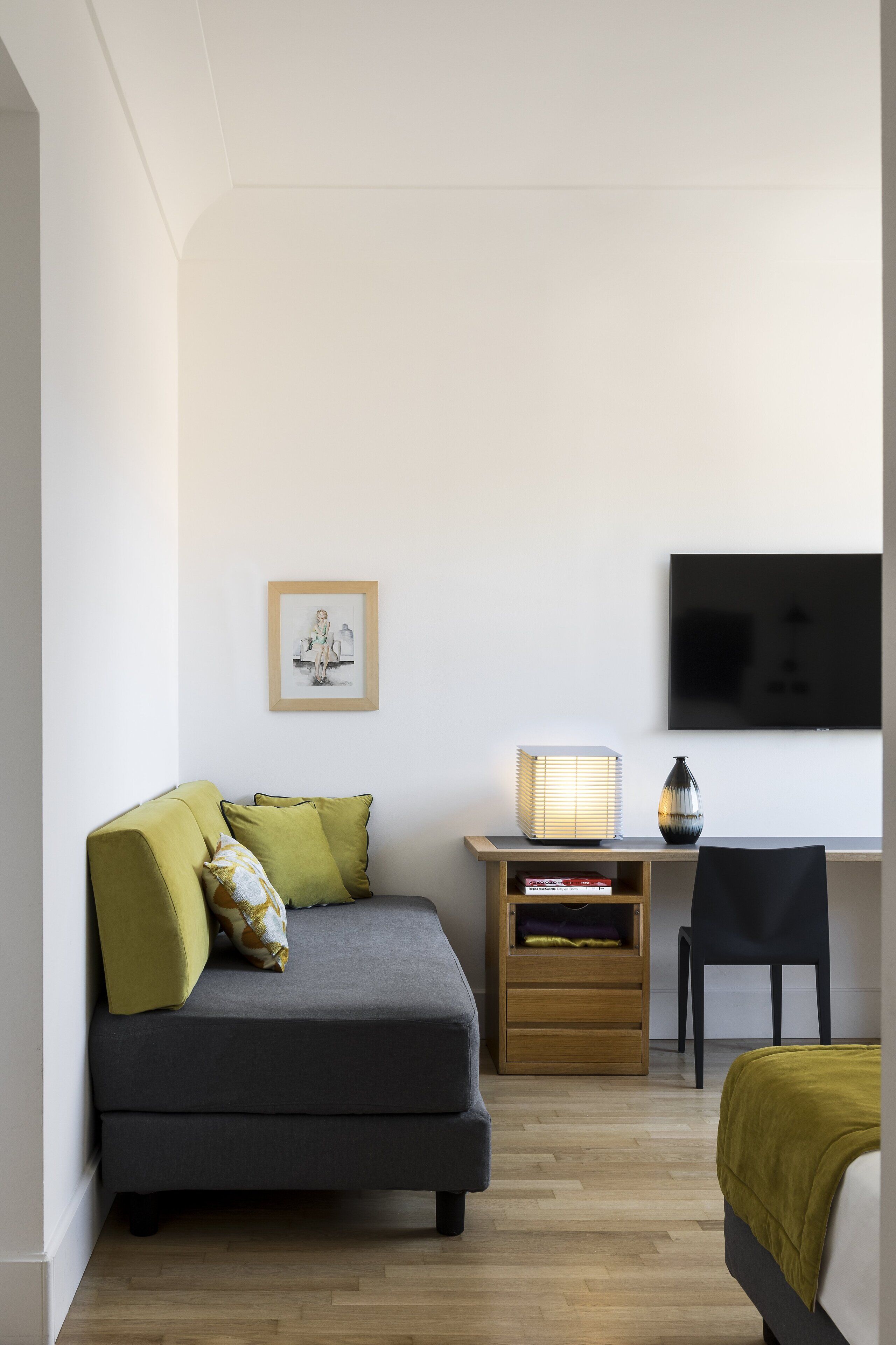 Un dormitorio moderno y minimalista con un sofá gris, una pequeña mesita de madera, una silla negra y una cama con una manta verde.